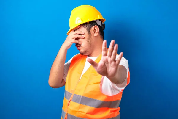 Fat asian construction worker man wearing uniform and helmet over isolated blue background covering eyes with hands and doing stop gesture with sad and fear expression. Embarrassed and negative concept.