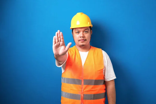 Fat asian workman wearing orange safety vest and yellow helmet making stop gesture with his hand denying a situation that could wrong.