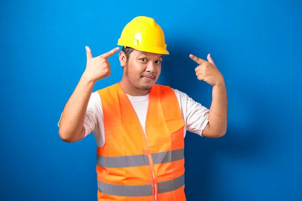 Asian construction worker man wearing orange safety vest and helmet over blue background smiling pointing to head with both hand finger, great idea or thought, good memory