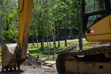 Toprak bir yolun kenarında bir kazıcı park halindedir. Yakından bakıldığında, çakıl taşlarına bastıran metal küreği görülür. Ağaçlar temizlenmeden önce sarı inşaat makinesi görülür..