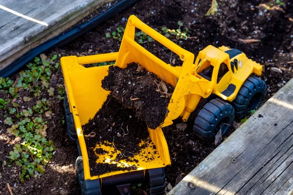 黄色いおもちゃのホイールローダーは 庭の耕運機で同じようなおもちゃのダンプトラックの後ろに土を入れるバケツをアンロードしています — ストック写真