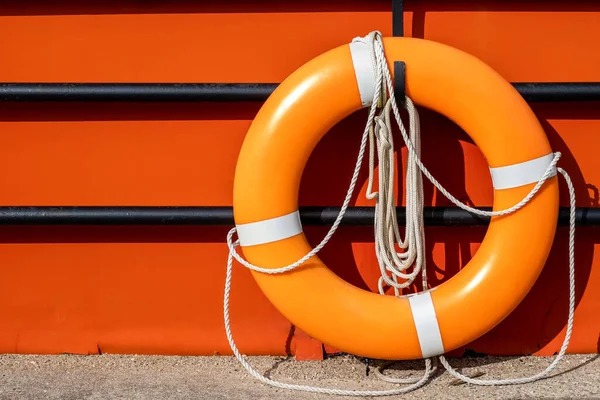 An orange life ring hangs on a hook on a black fence in front of an orange background surface. The life preserver has white ropes attached.