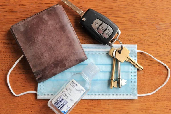 Flat lay of car keys, brown leather wallet, face mask and hand disinfectant gel