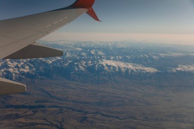 Uçuş sırasında uçağın lombozundan çıkan karlı dağlar. Güzelliğin panoramik görüntüsü yükseklikten. Yolculuk ve ulaşım kavramı