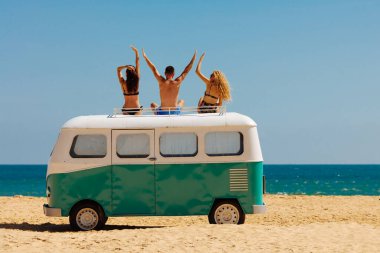 Sanddy Beach 'te hippi arabasının çatısında mayo giymiş üç mutlu turistin arkası görünüyor. Erkek ve bayan arkadaşlar Asya adasında dinleniyor ve güzel mavi deniz manzarasının tadını çıkarıyorlar..