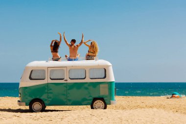 İki kadın ve bir erkeğin mavi ve beyaz retro otobüsün tepesinde oturup turkuaz denize baktığı arka plan görüntüsü. Mutlu arkadaşlar tatil boyunca kumsalda güneşli günlerin tadını çıkarıyorlar..