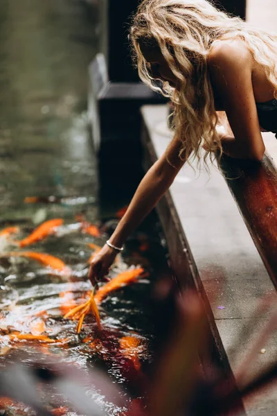 Side view of beautiful woman with curly hair in summer clothing standing on knees near edge of pond full of exotic fishes. Happy female tourist playing with marine animals using tropical flower