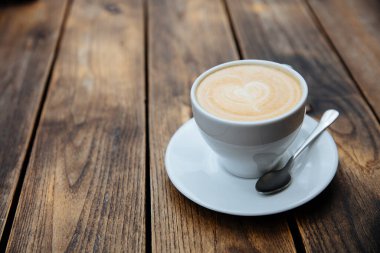 Beyaz seramik bir fincan taze demlenmiş kahve ve kalp şeklinde latte sanatı. Parlak kaşıkla servis edilen sıcak bir içecek. Koyu tahta arkaplan.