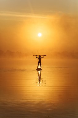 Yüzme şortlu güçlü adamın silueti gölün üzerinde güçlü bir sabah sisiyle kürek çekiyor. Su aktivitesi ve açık hava eğlencesi kavramı.