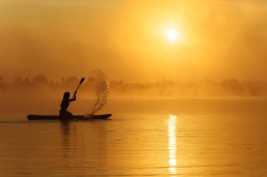 Gölde yaz gündoğumu sırasında yemek güvertesinde aktif erkek raket eğitimi. Kürekten su fışkırıyor. Siluetteki kişi. Her yer sisli..