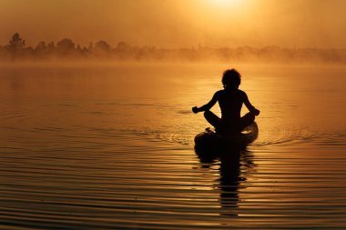 Gölün yukarısında güzel bir gündoğumu sırasında silüet giymiş kaslı genç adam sörf tahtasında meditasyon yapıyor. Etrafta yoğun sis var. Rahatlama ve sakinlik kavramı.