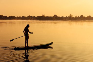 Gölde sabah vakti yemek masasında yüzmek için uzun kürek kullanan güçlü sporcu silueti. Su aktivitesi ve sağlıklı yaşam tarzı kavramı.