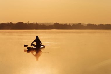 Silüetteki kaslı sporcu, sabahları yemek masasında yüzmek için uzun kürek kullanıyor. Gölde ağır sis arasında en sevdiği hobiyi yapan sporcu..