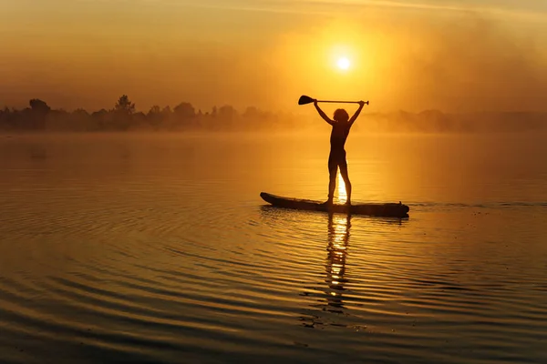Silhouette of young man with athletic body standing up on sup board and holding paddle above head. Amazing sunrise on background. Concept of water sport.