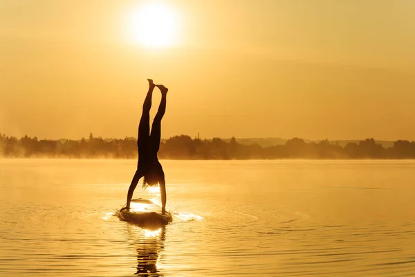 Strong fit guy standing on hands and keeping legs straight in air while swimming on sup board. Young man in silhouette balancing on water during amazing sunrise.