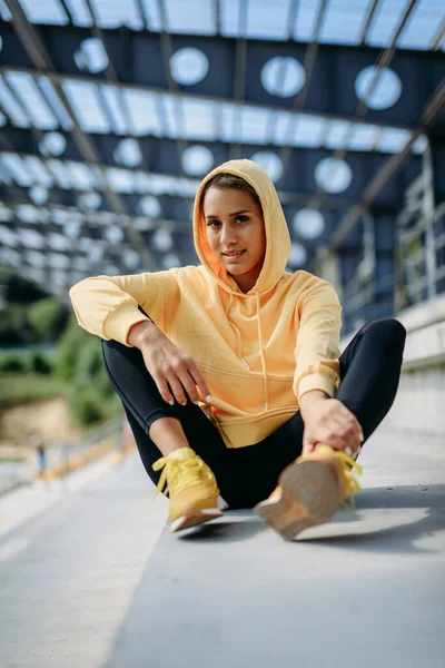 Charming young woman with slender body taking break outdoors after hard workout. Fitness female wearing black leggings and yellow sweater with hood on head. Sport concept.