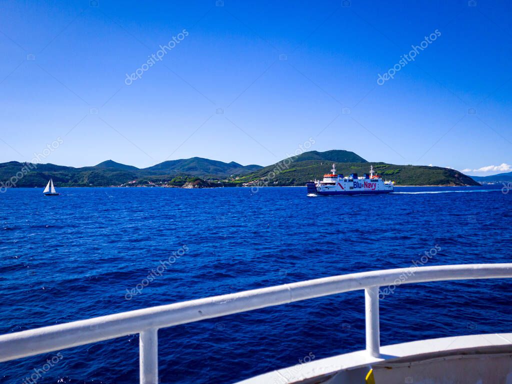 The ferry Acciarello of Shipping line Blu Navy at the Mediterranean see. In the background - white sailboat and island Elba. September 14, 2024. Portoferraio, Italy.