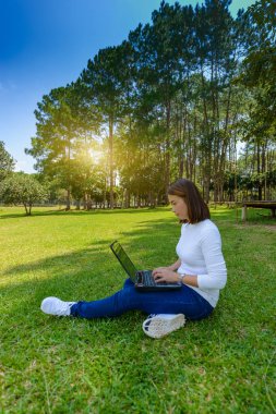 Dizüstü bilgisayar kullanırken yaz gün boyunca bağdaş ile Park yeşil çimenlerin üzerinde oturan genç ve güzel kadın portresi. arka plan manzara çam ormanının nedir