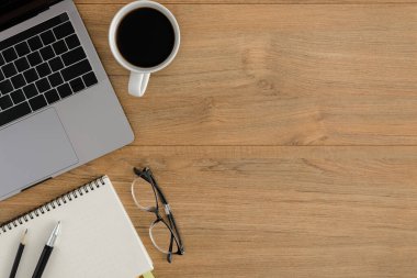 Düz yatıyordu, ahşap ofis masasının üstünde. Çalışma alanı boş defter, dizüstü bilgisayar, kalem, kahve fincanı ofis malzemeleri ve ahşap masa arkaplanı.