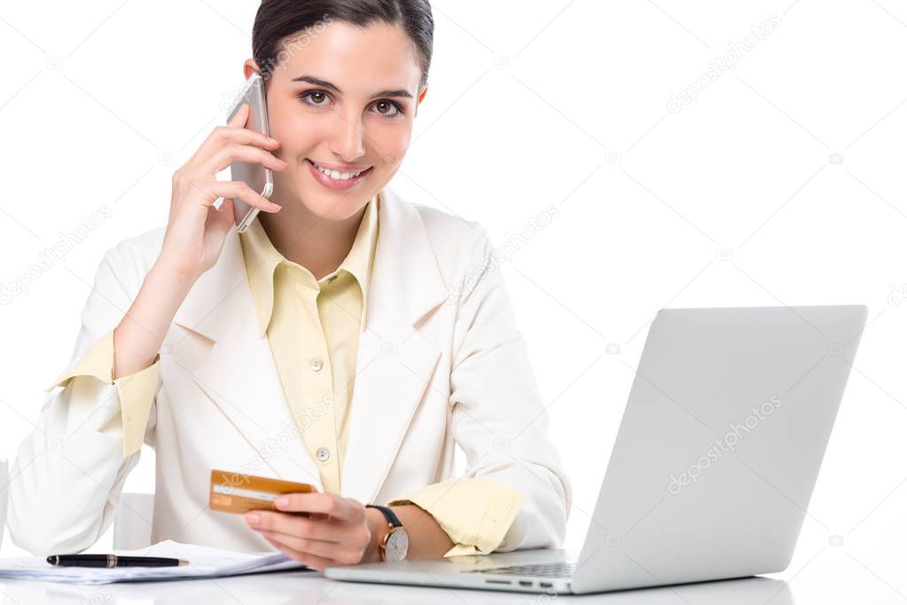 Beautiful smiling business woman showing credit card to camera while working with smartphone and laptop on her desk; Isolate on white background