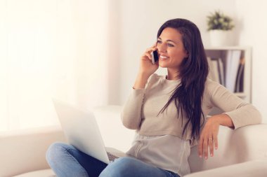Dizüstü bilgisayar kullanan bir kanepede oturan telefonda konuşurken gülümseyen kadın portresi 