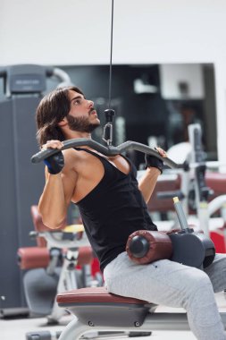 Uygun yetişkin vücut geliştirmeci çekerek bar kırmızı minder koltukta otururken halter makineye kablo ile bağlı