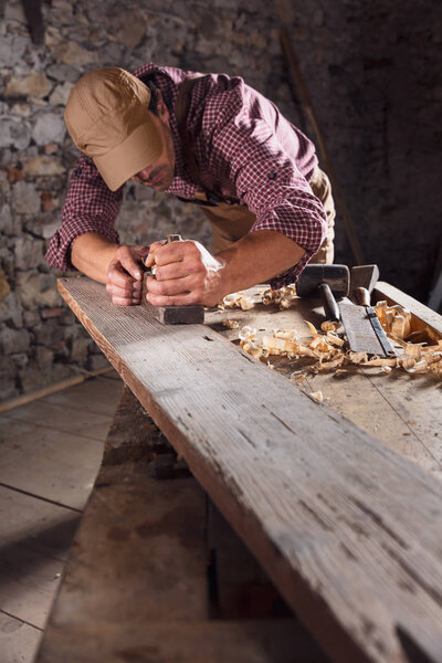 Carpenter smoothing out long wooden beam on top of table with hand plane and other planks in front of stone wall