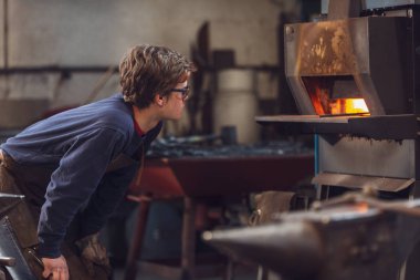 Genç blaksmith veya metalworker veya blaksmith bir metal işleme atölye bir yakma fırını ön planda bir örs ile izlerken