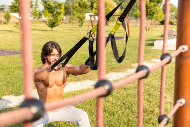 Güneşli bir yaz gününde parkta antrenman yaparken süspansiyon eğitmeniyle çömelme egzersizi yapan zinde bir genç; trx süspansiyon eğitim konsepti