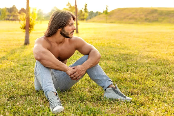 公園の緑の草の上に屋外に座っている間 強い腕とクールな態度で見てハンサムなひげの若い男 — ストック写真