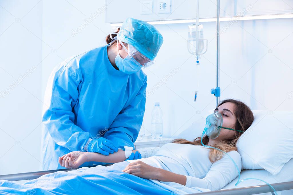 Nurse checking a intravenus drip on an ill patient wearing a positive pressure oxygen mask in hospital during the Covid-19 pandemic