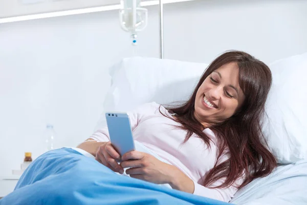 笑顔の女性患者が病院のベッドに横になり 彼のスマートフォンとチャット — ストック写真