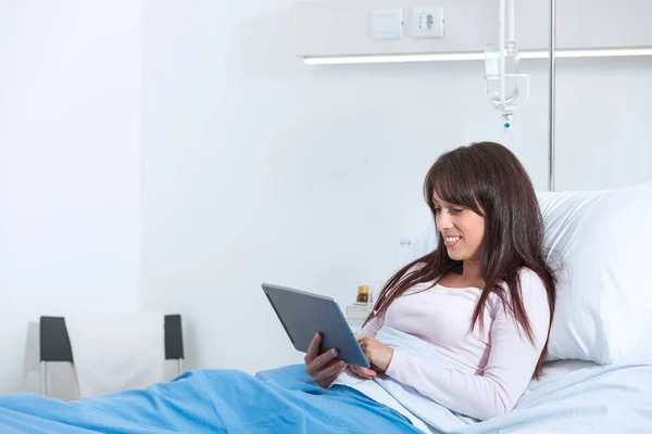 Young smiling patient lying in a hospital bed and she is using her digital tablet