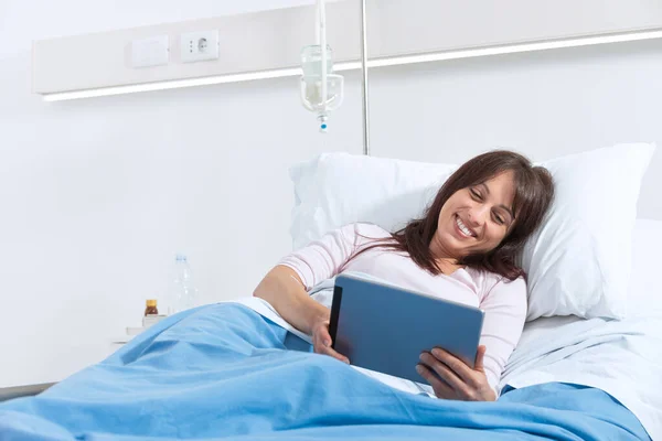 若いです笑顔患者は病院のベッドに横たわって 彼女はデジタルテーブルでビデオを見ています — ストック写真