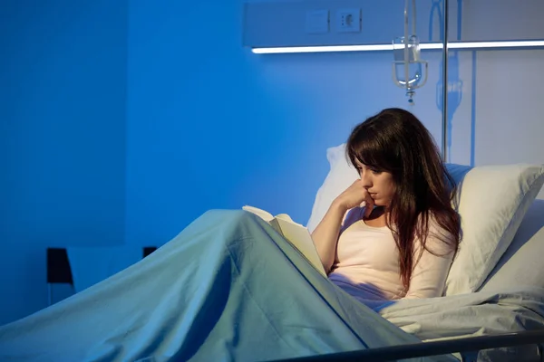夜に病院のベッドに横になって本を読んでいる若い女性 — ストック写真