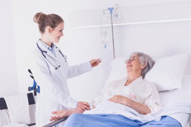 Hemşire ya da doktor, koğuşta yatan yaşlı bir kadının serumunu kontrol ediyor. Sıcacık bir gülümsemeyle sağlık hizmetleri konseptinde.