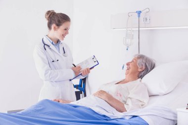 Doktor ya da hemşire, hastane koğuşlarında üst düzey bir hastayla sohbet ederken birbirlerine gülümserlerken notlar alıyorlar.