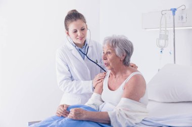 Yaşlı bir kadını dinleyen kadın doktor. Akciğerleri ve kalbi steteskopla hastahane koğuşunda tedavi görürken yatağında oturuyor.