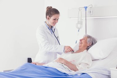 Gülümseyen kadın doktor koğuşta yaşlı bir bayanı muayene ederken steteskopla kalbini ve ciğerlerini dinliyor.