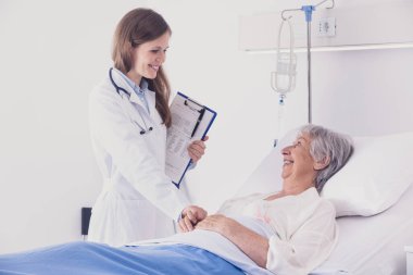 Gülümseyen kadın doktor ya da hemşire yaşlı bir hastayı yatağında rahatlatıyor. Hastane koğuşlarında elini tutmak için uzanıyor.