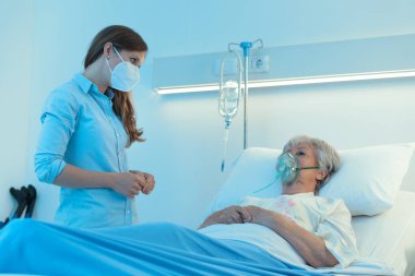 Hasta bakıcı ya da genç kız cerrahi maske takıyor. Morfin salgını sırasında hastane yatağında nefes almasına yardımcı olmak için pozitif basınçlı oksijen maskesi takan yaşlı bir kadını kontrol ediyor.