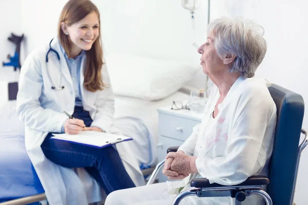 看護師又は医師が 車椅子に座っている笑顔の高齢者の女性のために病棟に患者ノートを書きます医療と医療の概念 — ストック写真