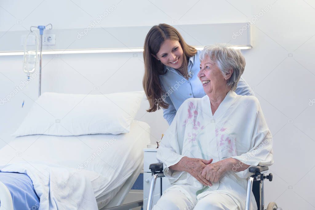 Carer or young daughter wheeling an elderly woman in a wheelchair bending down to smile at her indoors in a hospital room on a ward or old-age home