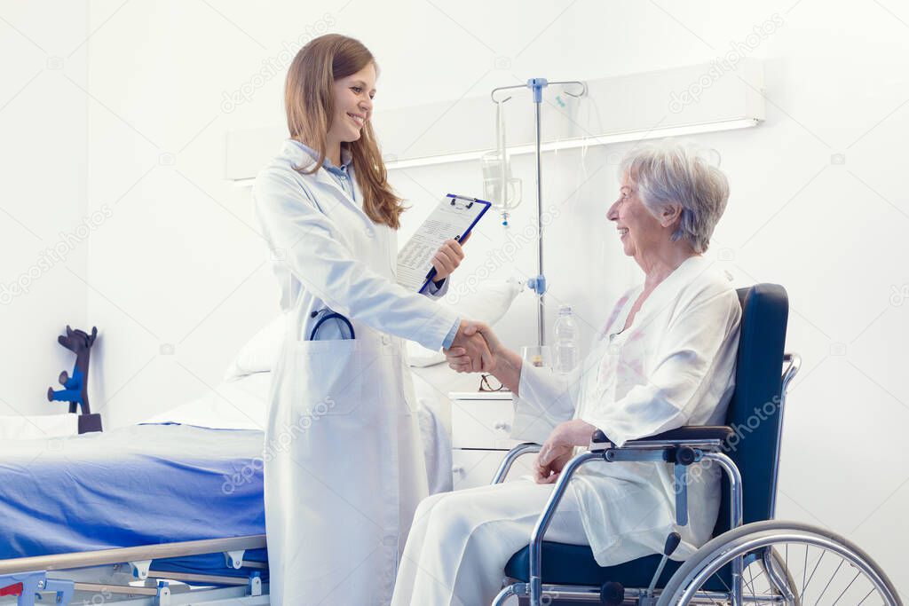 Smiling friendly woman doctor shaking hands with an elderly patient seated in a wheelchair in her room in the hospital during ward rounds