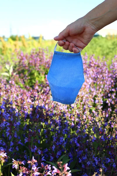 Hand holding cotton face mask at the nature flowers field background. Home made cloth face mask against coronavirus Covid 19.