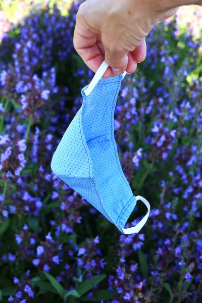 Hand holding cotton face mask at the nature flowers field background. Home made cloth face mask against coronavirus Covid 19.