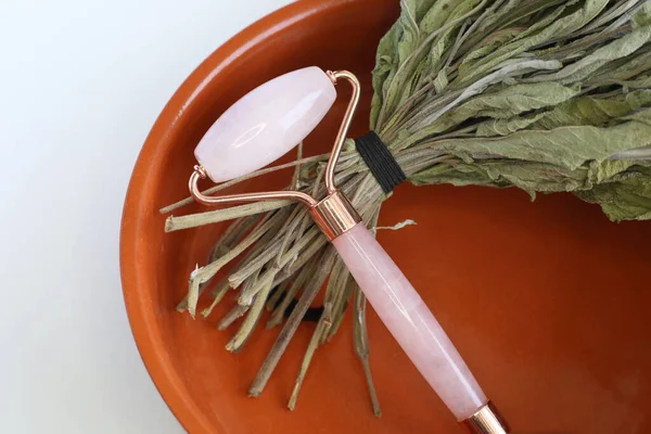 Pink face roller laying on dry herbs on white background. Beauty roller tools. Quartz roller massager for face.