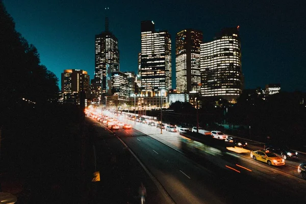 Auto-estrada com lotes de carros à noite com altos edifícios iluminados à noite no fundo — Fotografia de Stock