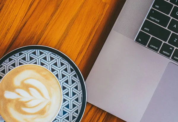 Primer plano de una taza de capuchino bellamente hecho al lado de un ordenador portátil en una superficie de madera — Foto de Stock