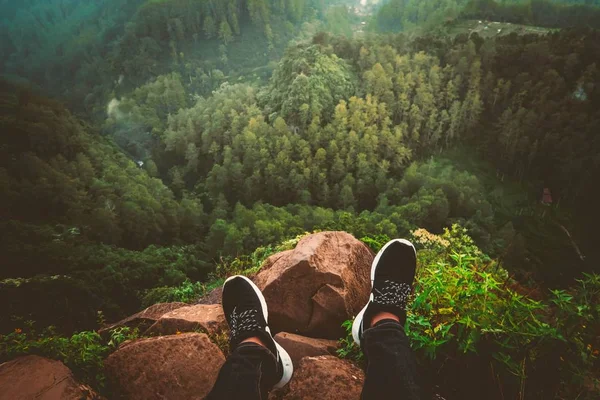 Los pies de la persona en zapatillas de deporte sentadas en un borde rocoso acantilado mirando a un hermoso bosque y colinas — Foto de Stock